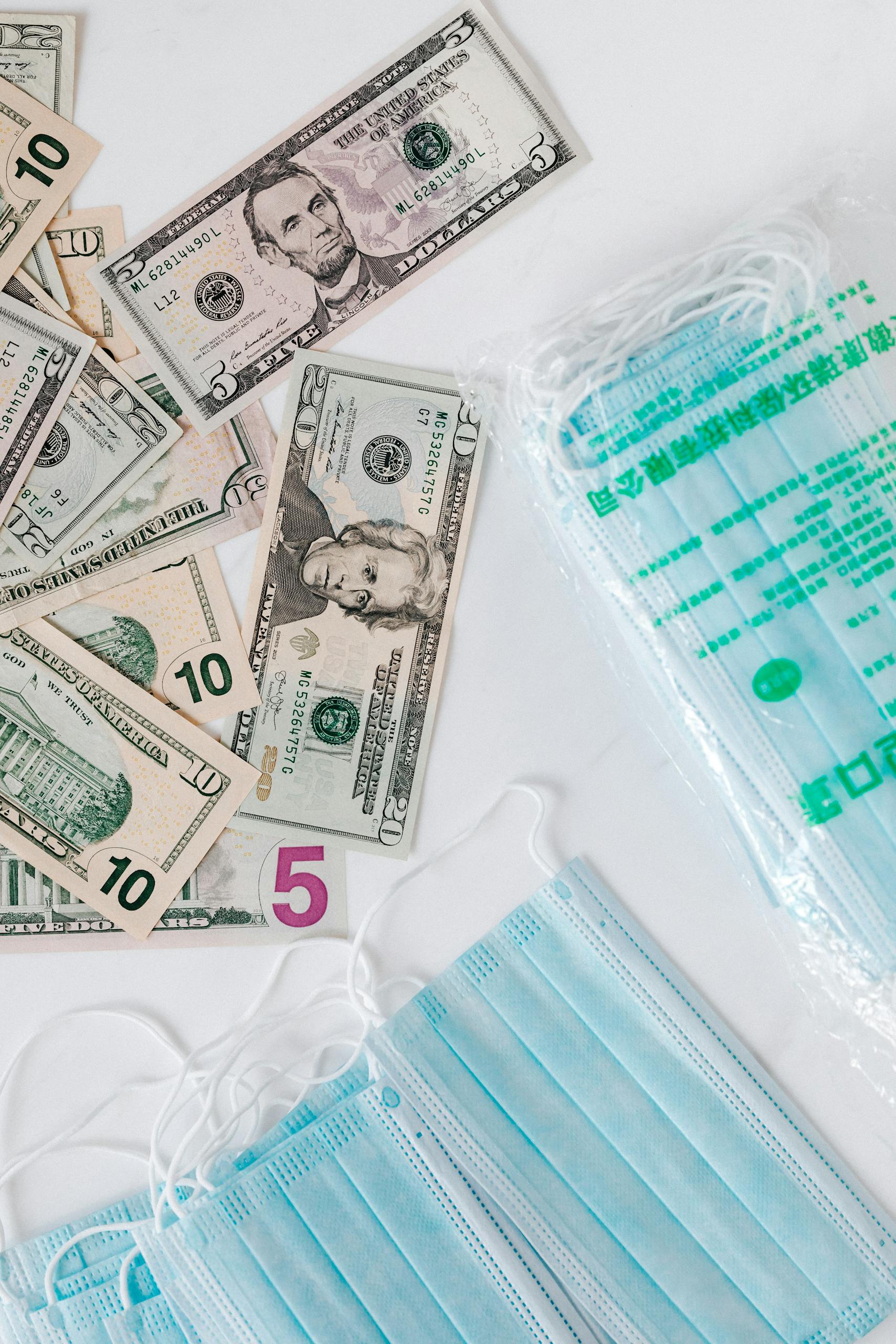Top view composition of disposable medical masks placed on white table near scattered randomly various paper banknotes of American dollars